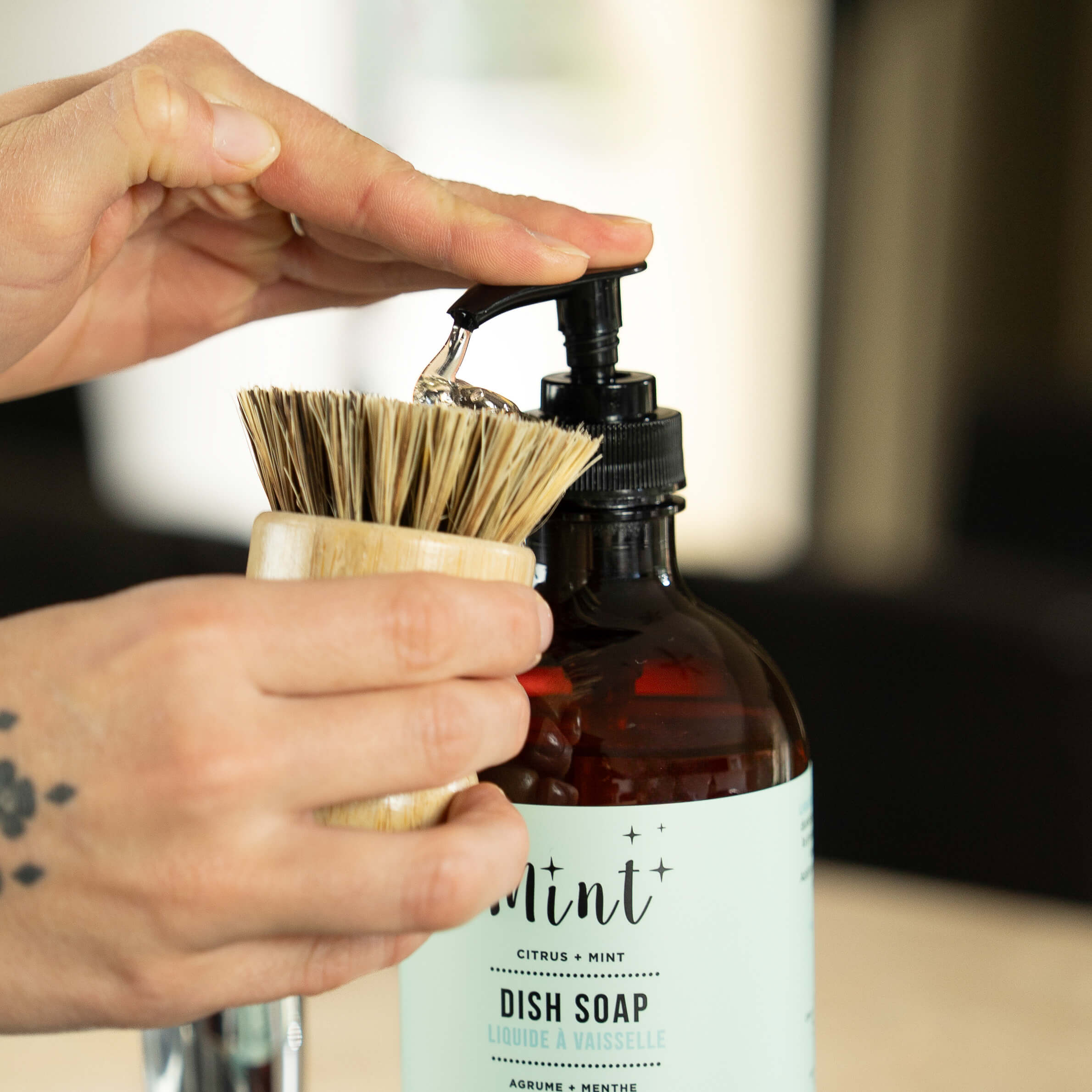Person holding a bottle of dish soap with a brush attached, against a blurred background.