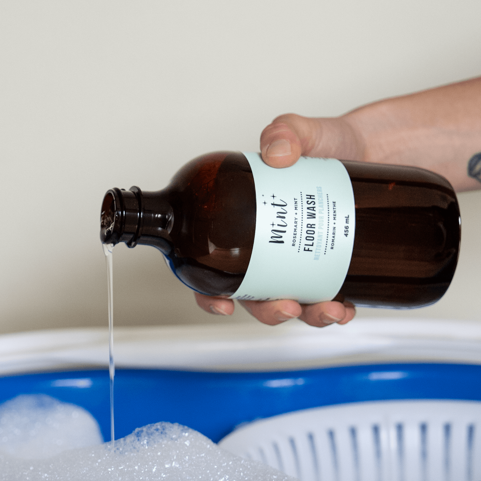Person pouring a liquid from a brown bottle labeled 'Mint' into a bathtub filled with bubbles.