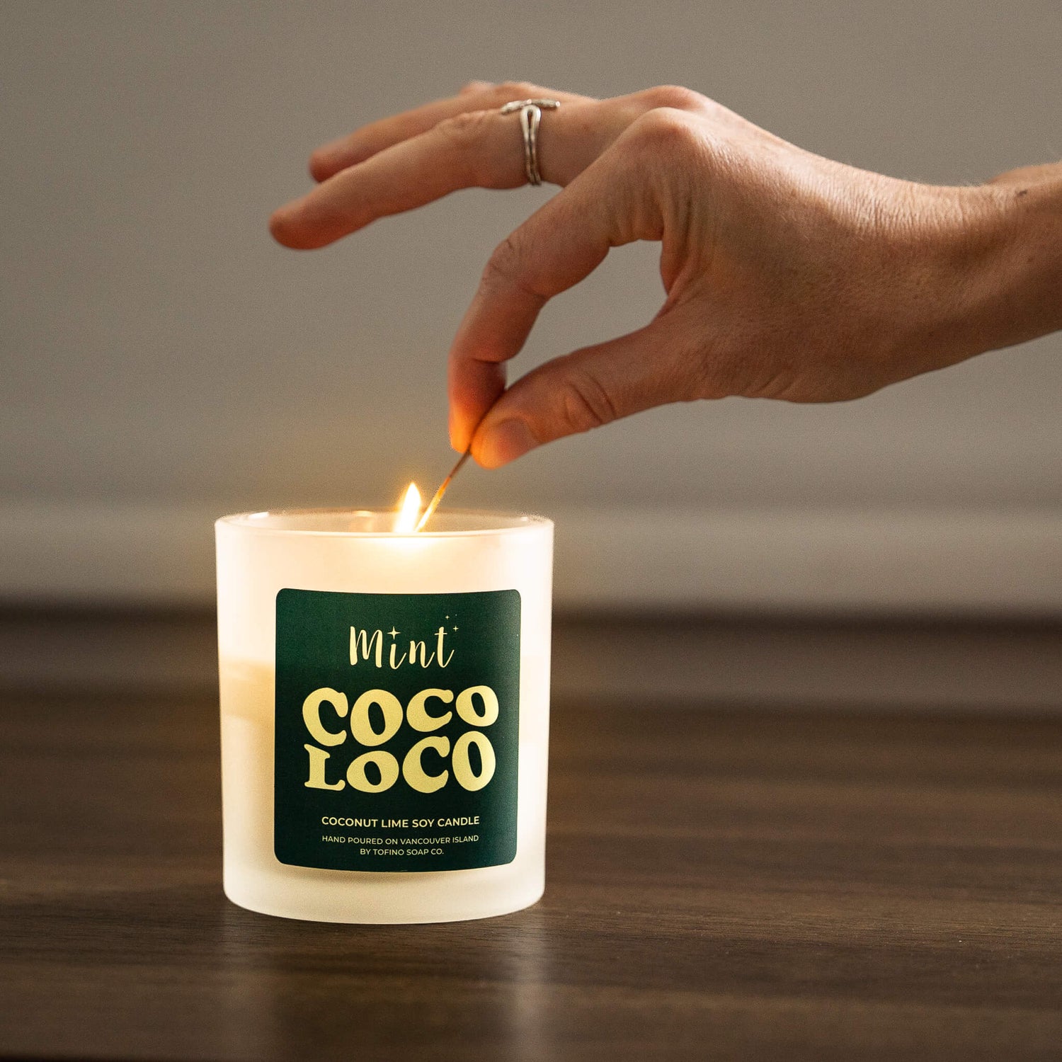 Hand lighting a Coco Loco candle with a green label on a wooden surface.