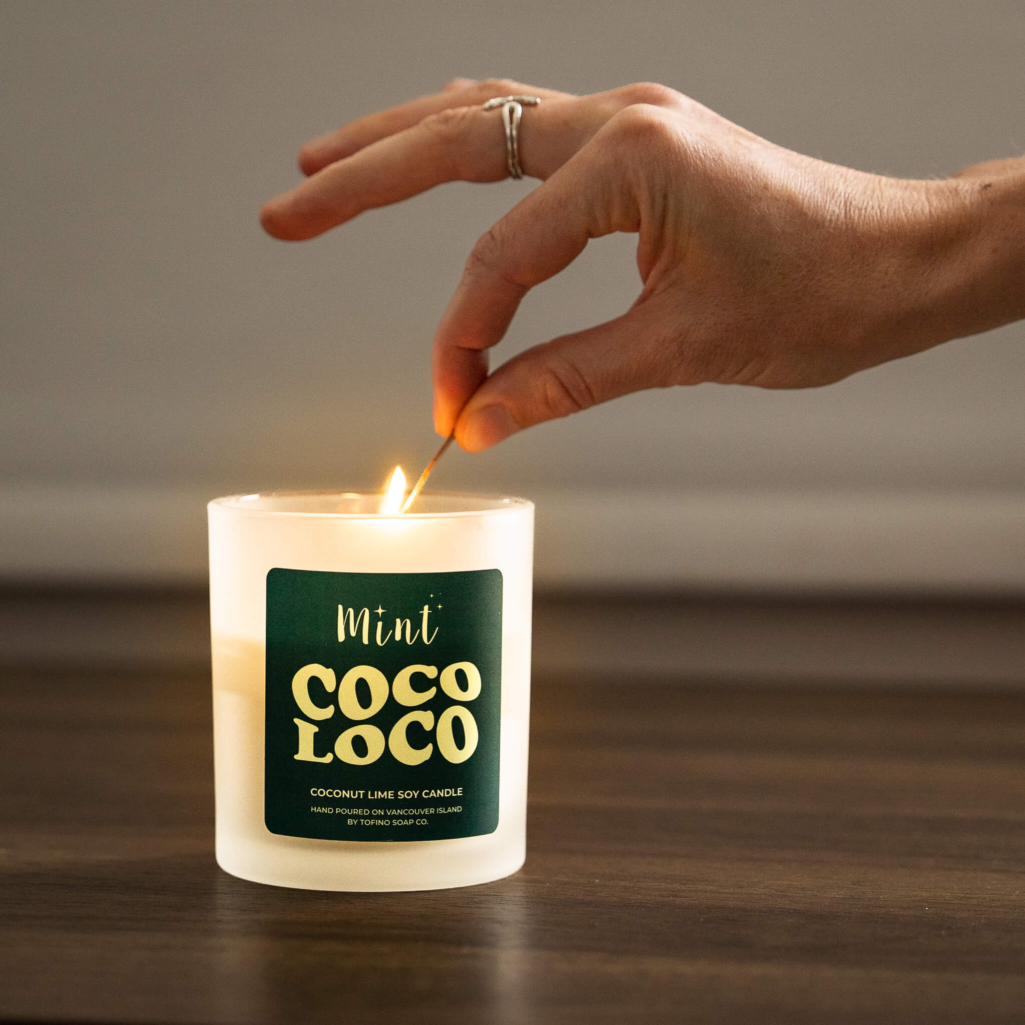 Hand lighting a Coco Loco candle with a green label on a wooden surface.
