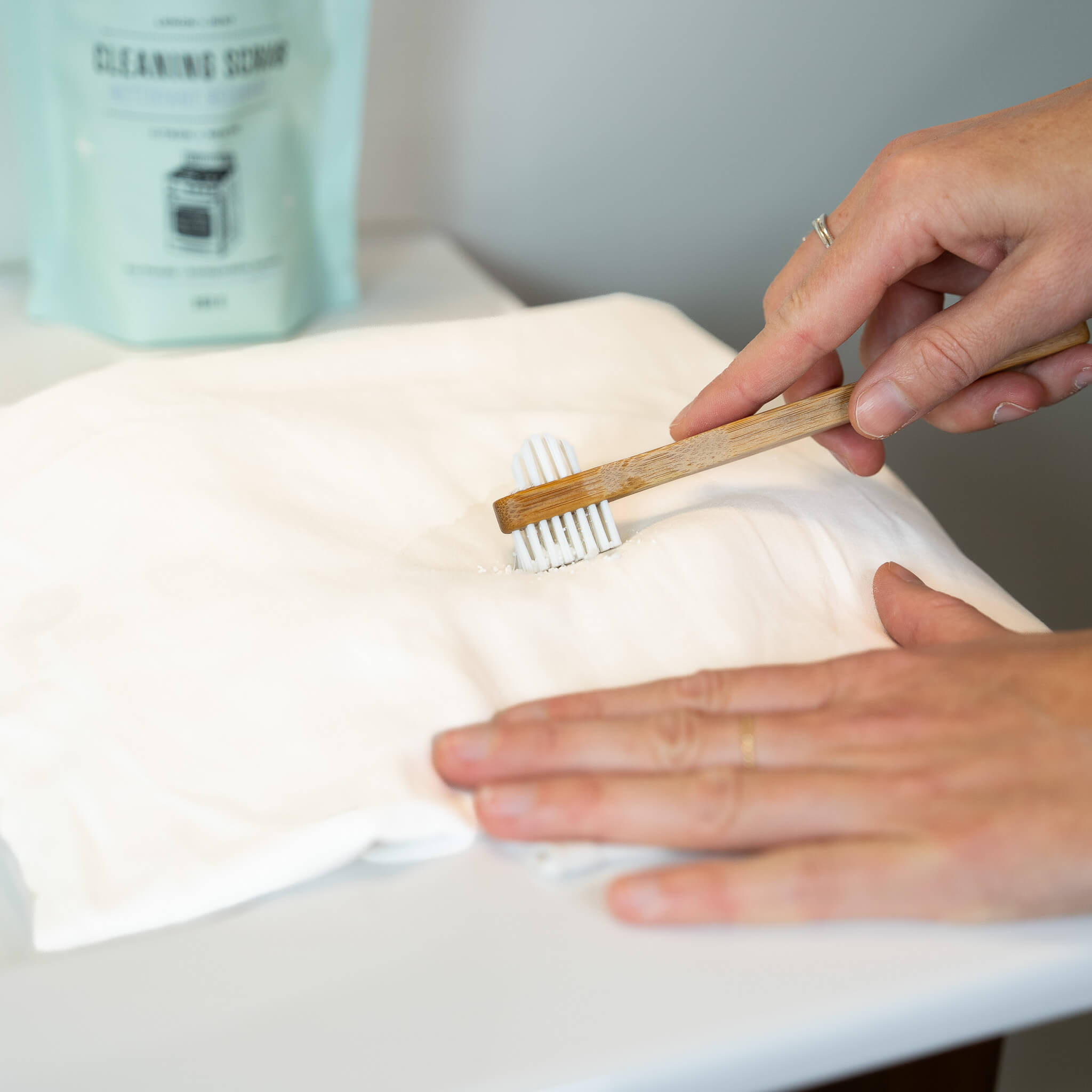 Person cleaning a white cloth with a wooden brush and a container of cleaning scrub in the background.