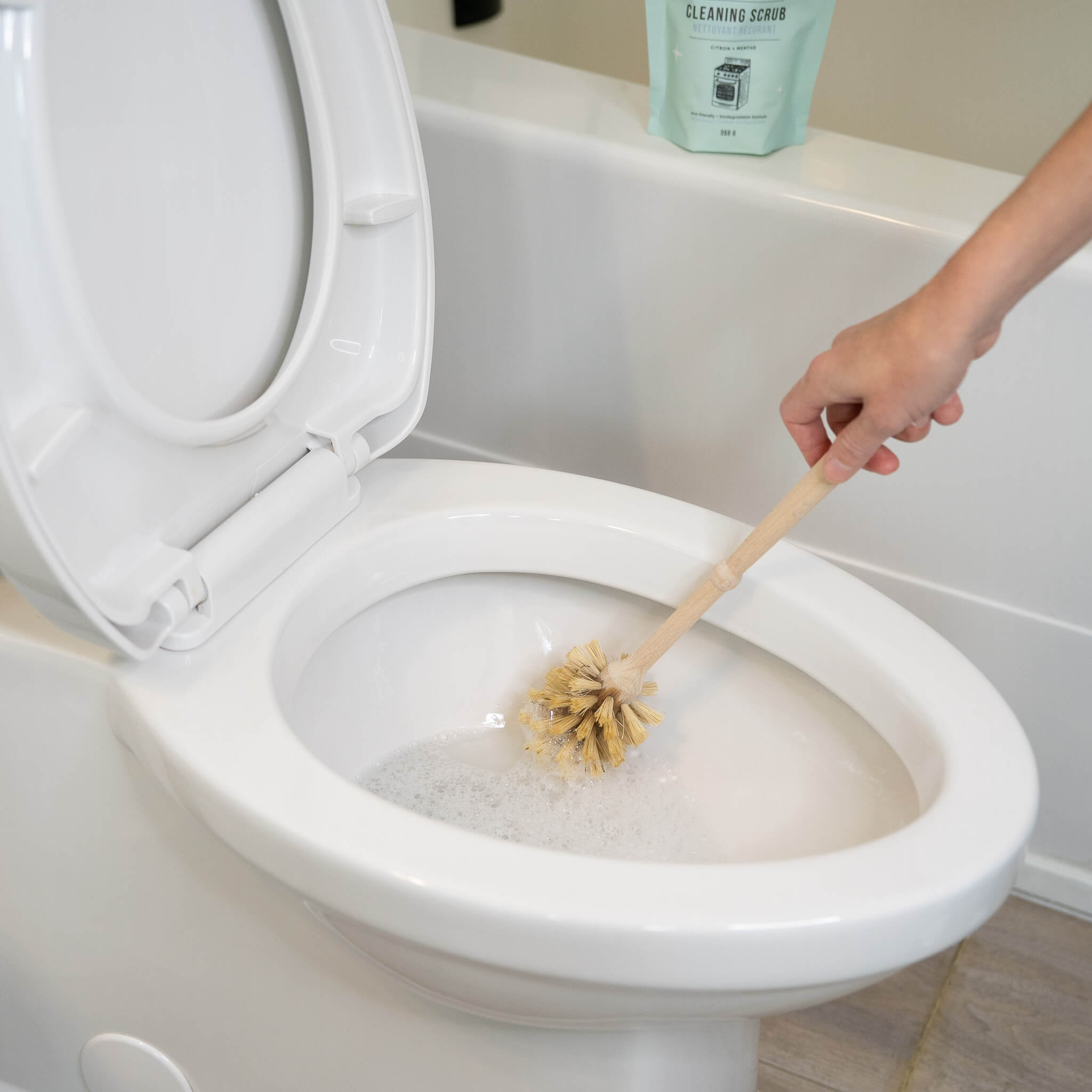 Person cleaning a toilet with a brush and cleaning scrub in the background.