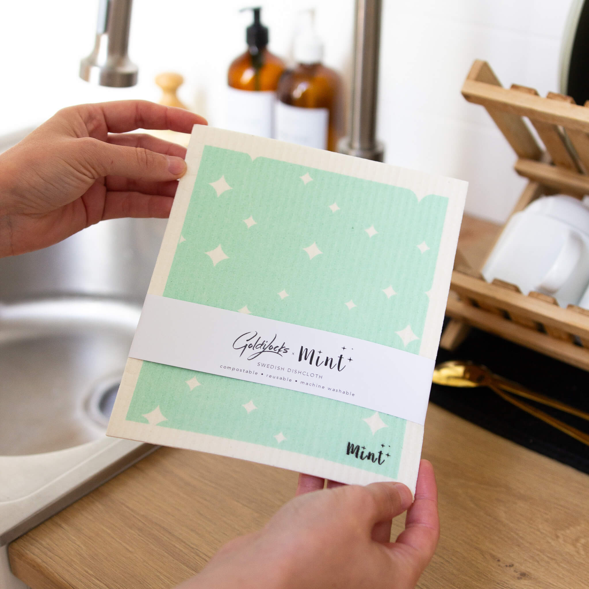 Hands holding a Mint x Goldilocks Swedish Dishcloth labelled over a kitchen sink, showcasing the mint green star pattern.