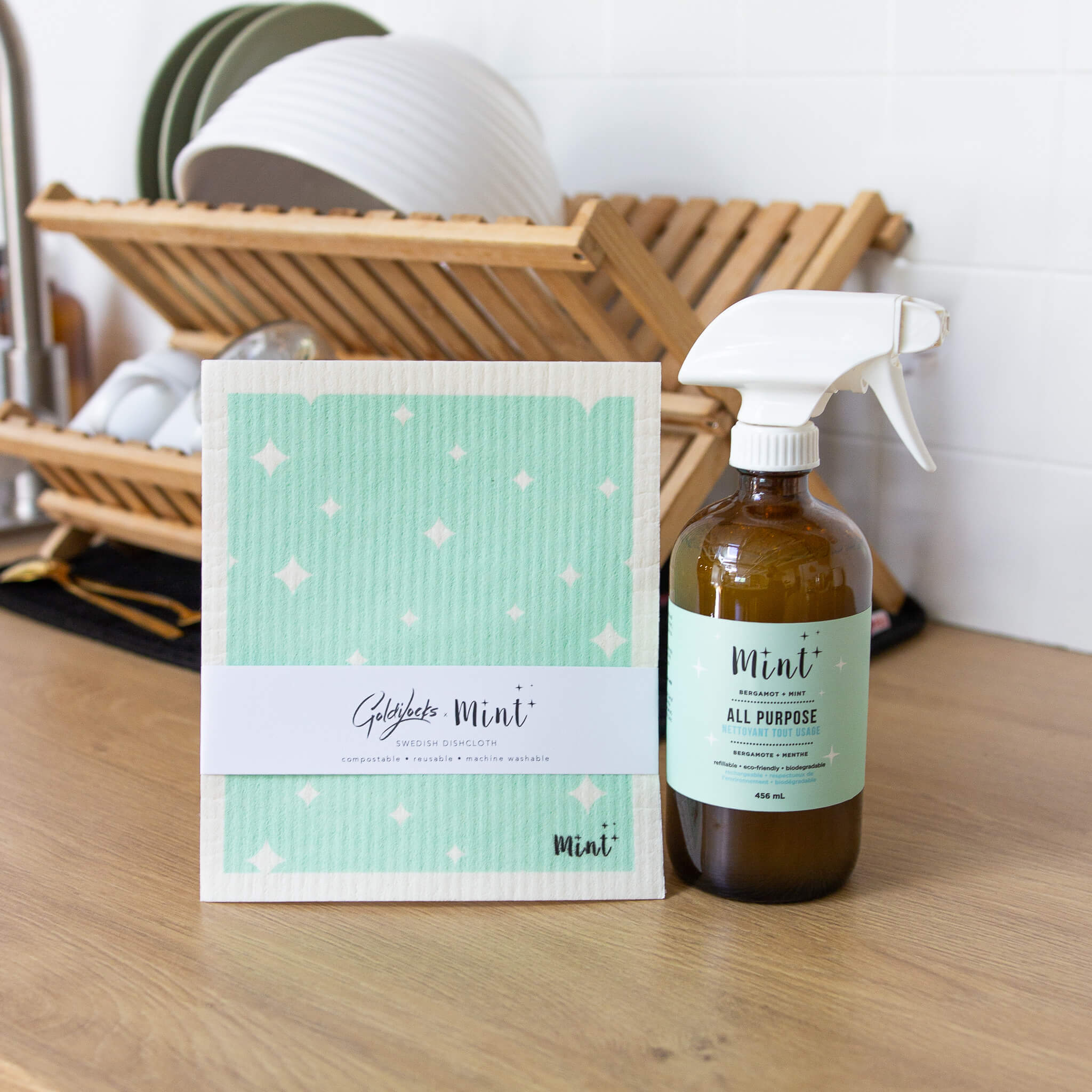 Mint x Goldilocks Swedish Dishcloth standing beside a Mint All Purpose Cleaner bottle on a kitchen counter with dish rack in the background.