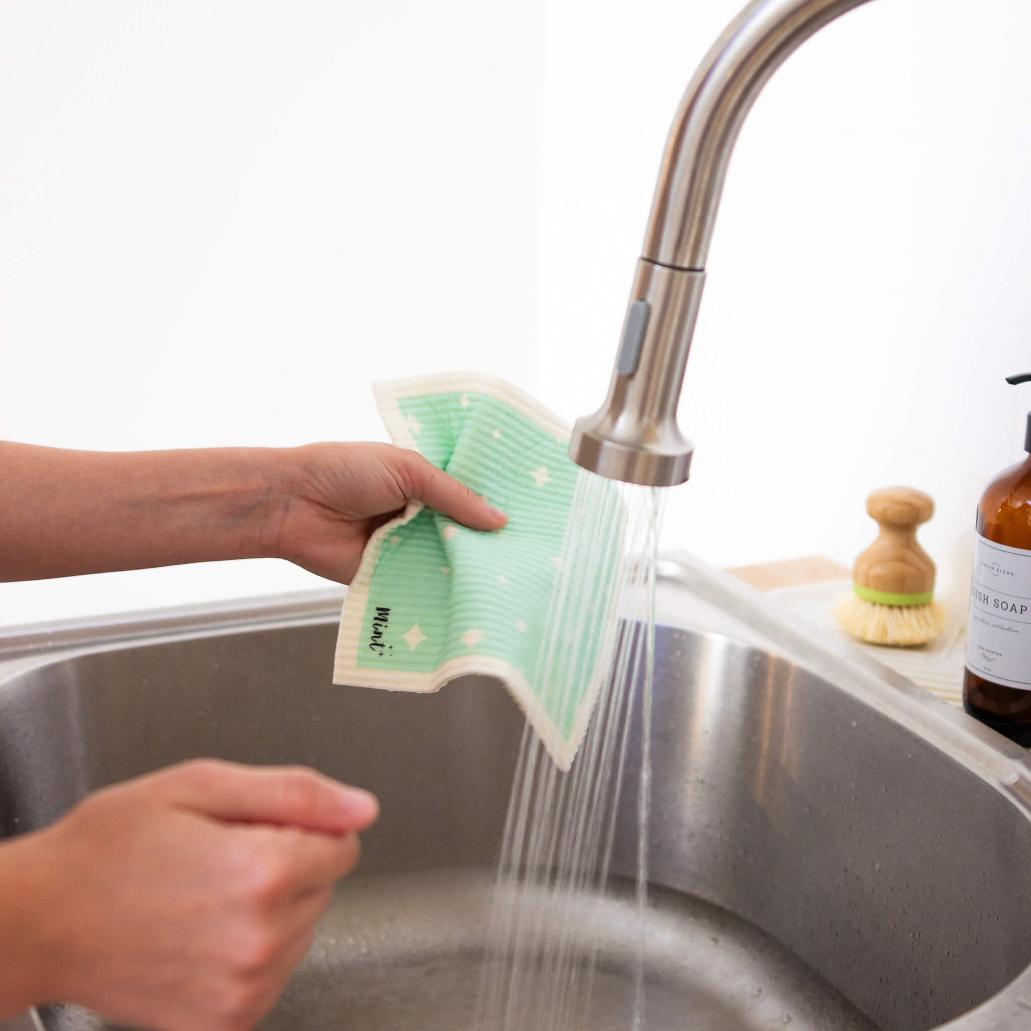 Person rinsing a Mint x Goldilocks Swedish Dishcloth under running tap water in a stainless steel kitchen sink.