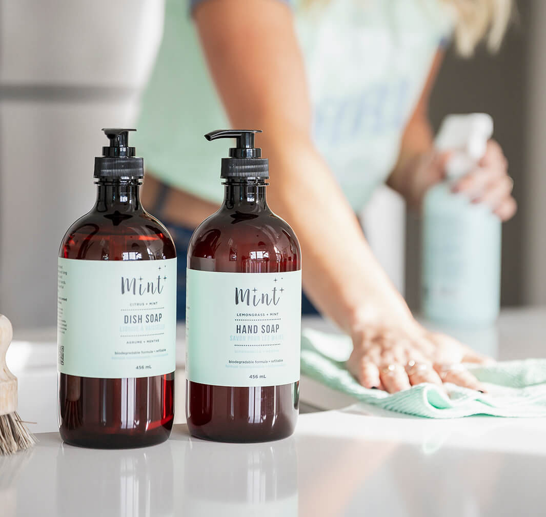 Two bottles of Mint dish soap and hand soap on a kitchen counter with a person cleaning in the background.