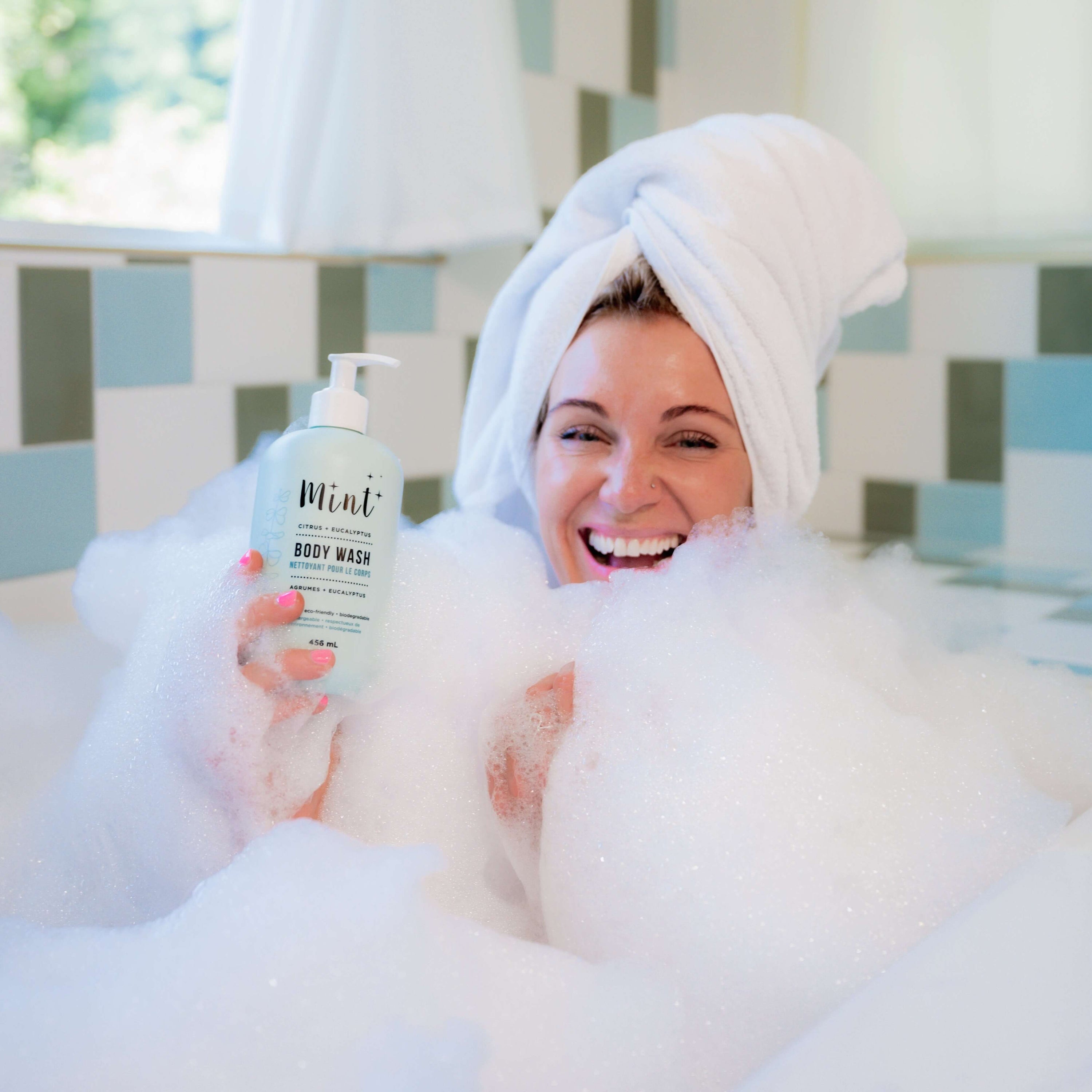 Monika, co-founder of Mint Cleaning Products, smiling and enjoying a bubble bath while holding a 456 mL bottle of "Mint+ Body Wash." She has a towel wrapped around her head, surrounded by fluffy bubbles. The light blue bottle features the "Citrus + Eucalyptus" scent and eco-friendly branding. The background shows a bright and inviting bathroom with a tiled wall and natural light streaming in.