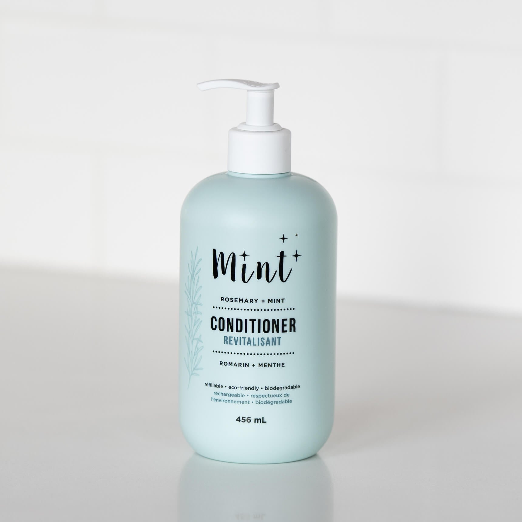 A 456 mL light blue bottle of "Mint+ Conditioner" with a pump dispenser, displayed on a white countertop. The label highlights the scent "Rosemary + Mint" and includes bilingual text, "Revitalisant" and "Romarin + Menthe." It emphasizes eco-friendly attributes such as being refillable, biodegradable, and sustainable. A subtle botanical illustration adorns the left side of the label, adding a natural element to the design. The background features a bright and minimalistic white setting.