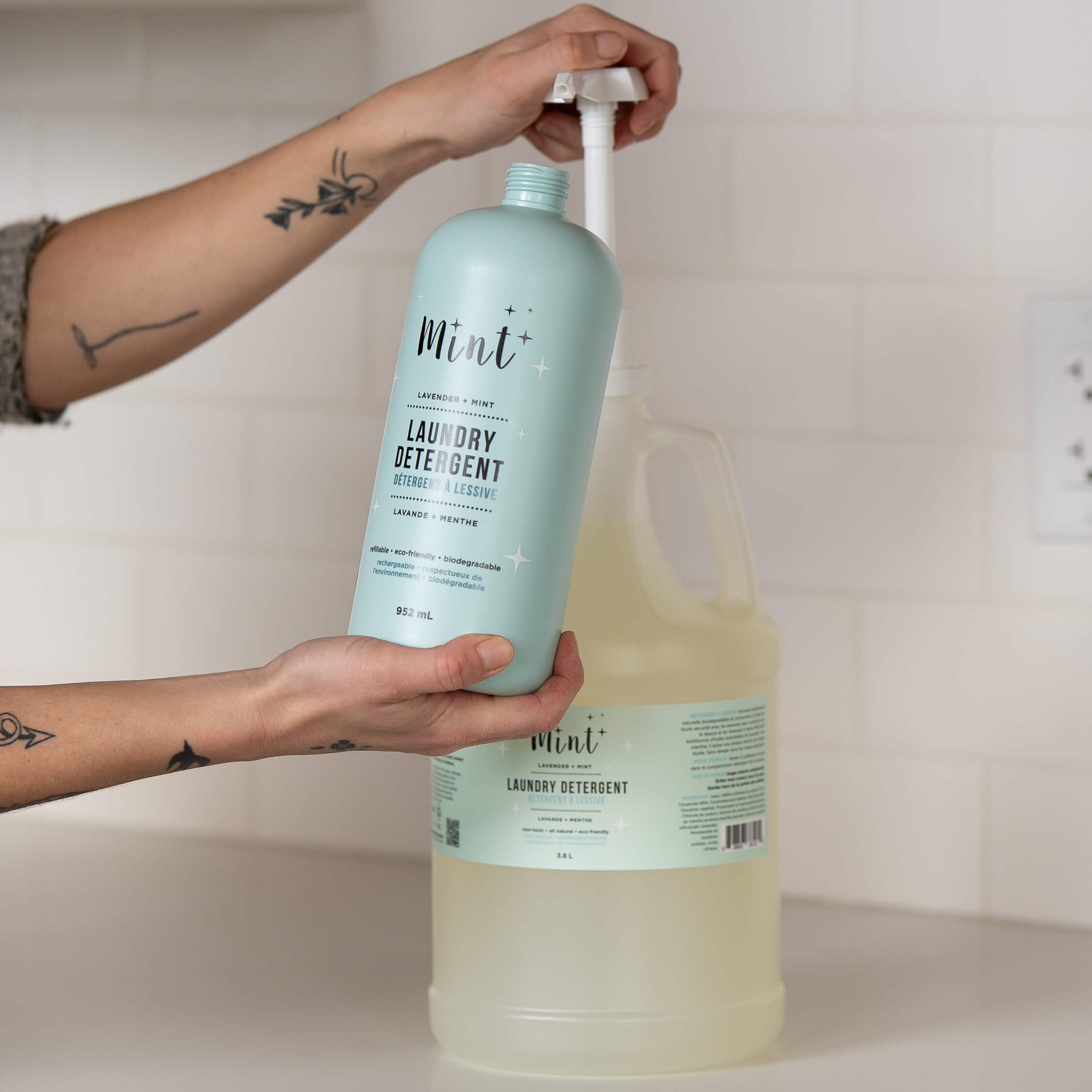 A person holding a 952 mL light blue bottle of "Mint+ Laundry Detergent" with the scent "Lavender + Mint," being refilled from a 3.8 L refill jug. The smaller bottle features bilingual text and eco-friendly attributes, such as being refillable and biodegradable. The person’s tattooed hands are visible, with the large jug in the background on a white countertop with a tiled wall.