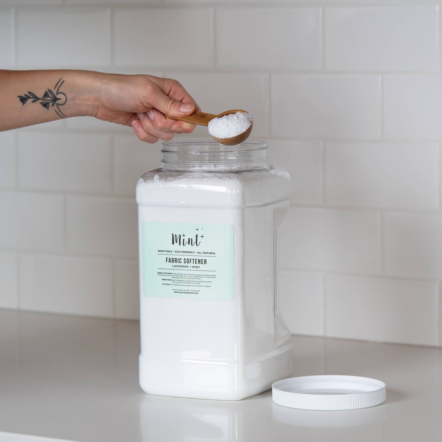 A large white container of "Mint+ Fabric Softener - Lavender + Mint" with its lid off, placed on a white countertop. A hand with a tattoo is holding a bamboo scoop with product over the open container, against a minimalist subway tile background.