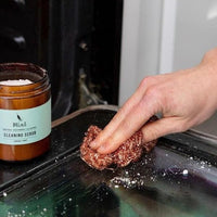 hand using copper scrub pad with Mint natural cleaning scrub powder to scrub glass inside oven door