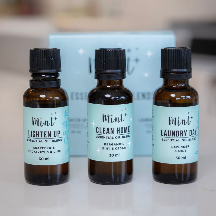 Three bottles of Mint essential oils, labeled 'Lighten Up,' 'Clean Home,' and 'Laundry Day,' placed on a countertop with a Mint packaging box in the background.