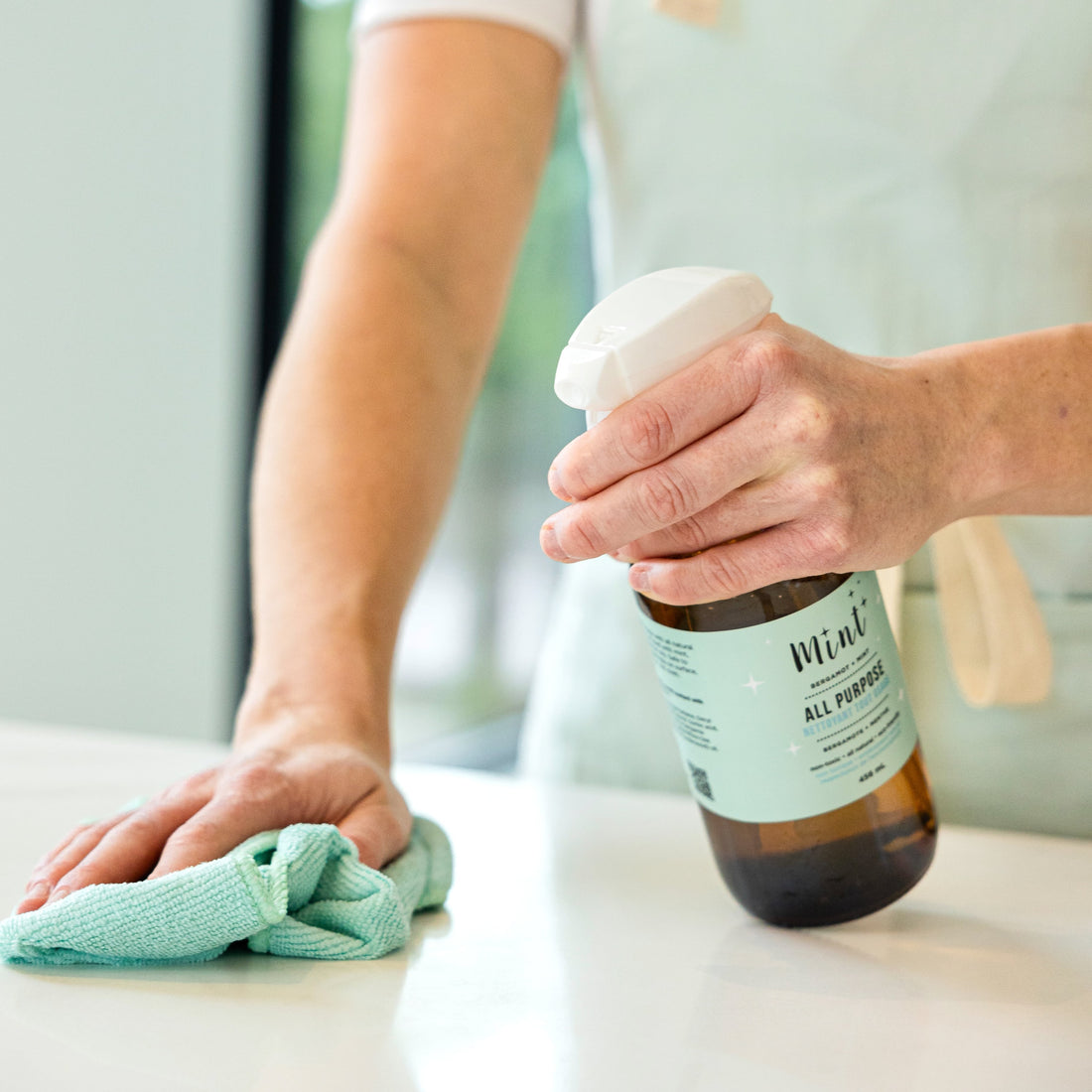 Person using Mint All Purpose Cleaner in a glass bottle with a spray nozzle, cleaning a white surface with a green cloth.