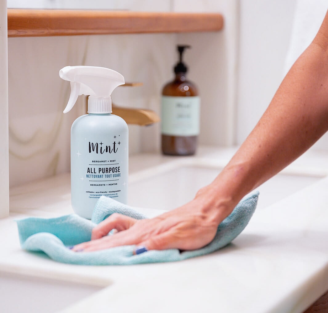 A hand wipes a countertop with a light blue cloth near a bottle of Mint All Purpose Cleaner, designed for versatile cleaning with eco-friendly ingredients, ideal for kitchens and bathrooms.