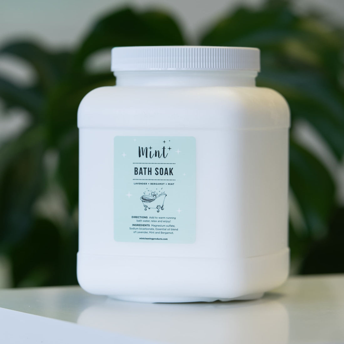 Mint Bath Soak refillable container in a minimalist design, placed against lush green foliage, highlighting its natural and eco-friendly qualities.