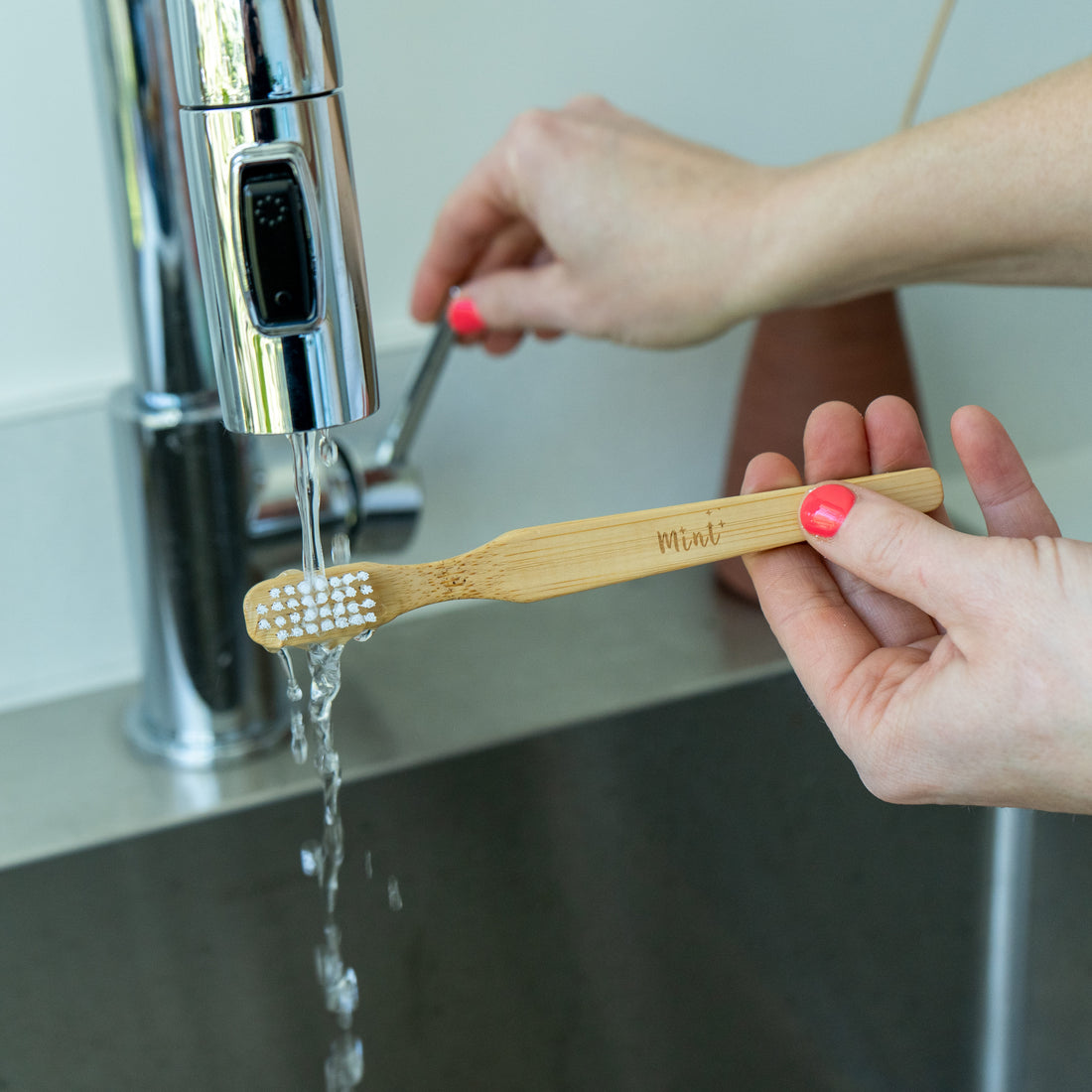 A bamboo cleaning brush with white bristles being rinsed under a stainless steel faucet, showing the Mint logo on the handle.