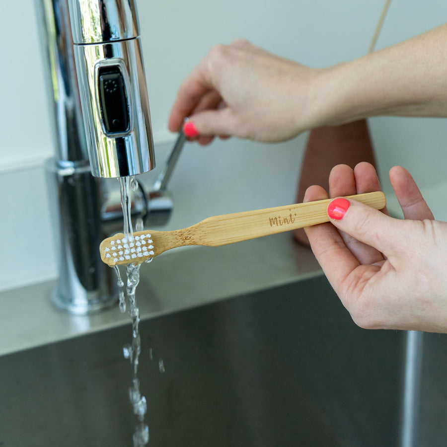 A bamboo cleaning brush with white bristles being rinsed under a stainless steel faucet, showing the Mint logo on the handle.