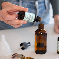 Person pouring Mint Clean Home Essential Oil Blend into an amber bottle. Tools for essential oil handling are on the table.