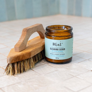 Mint Cleaning Scrub in a 250g amber glass jar, displayed on a tiled surface with a wooden scrubbing brush. The label highlights its natural and eco-friendly ingredients.