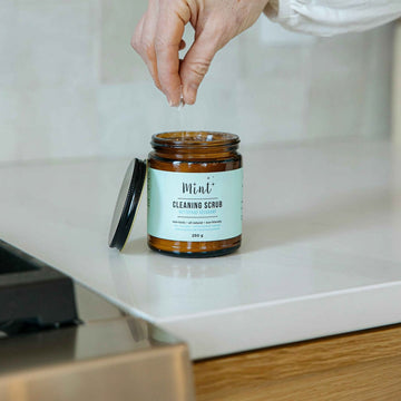 Mint Cleaning Scrub in a 250g amber glass jar, with a hand sprinkling the scrub on a white countertop. The label highlights its natural and eco-friendly ingredients.