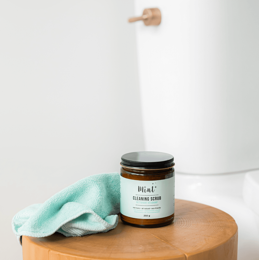 Mint Cleaning Scrub in a 250g amber glass jar, placed on a wooden stool next to a green cleaning cloth. The label highlights its natural and eco-friendly ingredients.