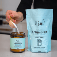 A person using a wooden scoop to transfer cleaning scrub from a Mint Cleaning Scrub 500g refill pouch into a 250g amber glass jar. The pouch is labeled with Lemon + Mint scent, highlighting its eco-friendly and biodegradable properties.