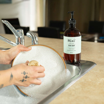 Person washing dishes with a bottle of Mint dish soap on a kitchen counter.