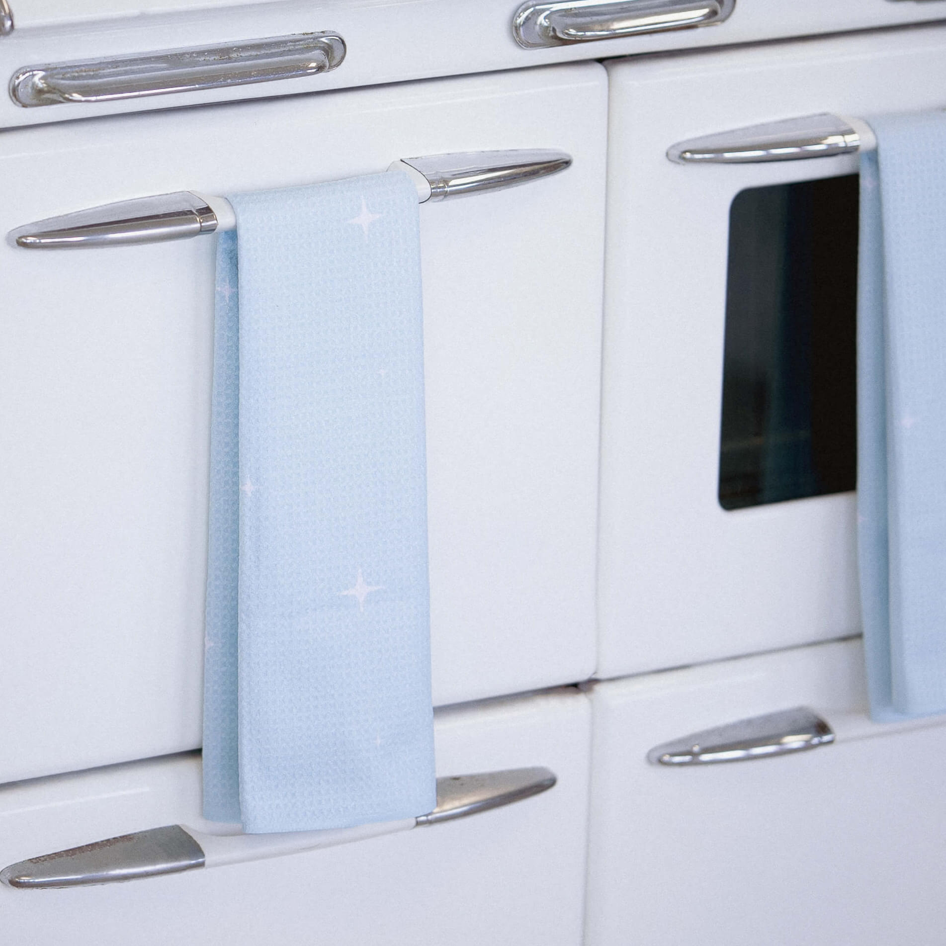 A light blue tea towel with a subtle star pattern hangs neatly on the handle of a vintage white oven with chrome accents. Each towel is made from 3.5 recycled plastic bottles and designed for both style and sustainability. Crafted from GeoWeave™ fabric, it offers quick-drying technology and superior absorbency—holding up to 7 times its weight.