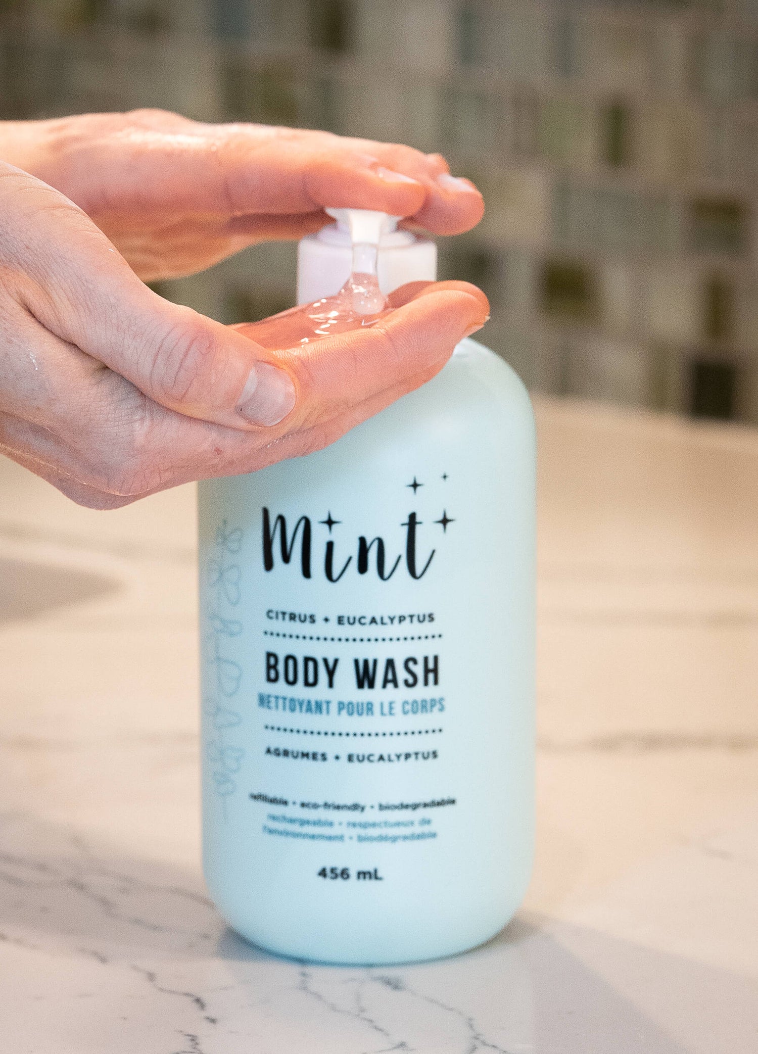 A close-up of a hand dispensing clear body wash from a light mint-green "Mint Citrus + Eucalyptus Body Wash" bottle. The bottle is placed on a white counter with a blurred background.