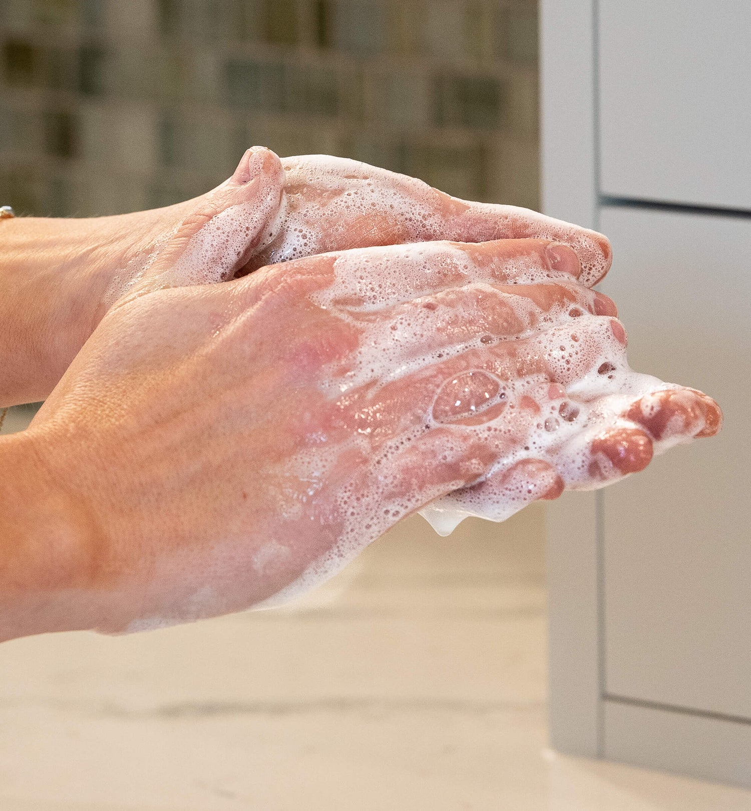 Hands lathered with white soap suds, captured in a close-up shot against a blurred tiled background.