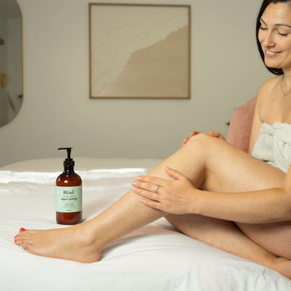Woman applying lotion to her leg with a bottle of lotion beside her on a bed.