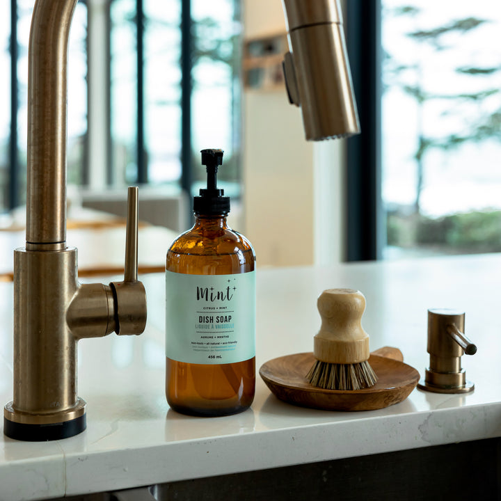 Mint Dish Soap in a 456ml amber glass bottle with a pump, placed on a kitchen counter next to a wooden dish brush and a brass faucet.