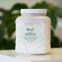 Mint Dishwasher Tabs in a refillable white container with green leaves in the background, emphasizing the natural and eco-friendly packaging.