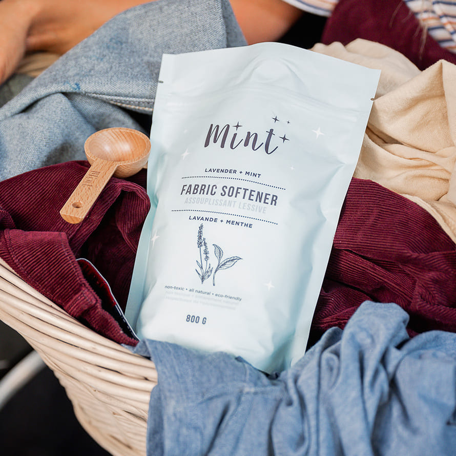 Mint lavender and mint fabric softener in an 800g refillable bag displayed in a laundry basket with assorted colourful clothes and a wooden scoop, emphasizing eco-friendly laundry care.