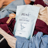 Mint lavender and mint fabric softener in an 800g refillable bag displayed in a laundry basket with assorted colourful clothes and a wooden scoop, emphasizing eco-friendly laundry care.