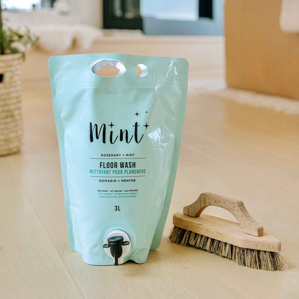 Mint Floor Wash in a 3L refill pouch with an easy-to-use spout, displayed next to a wooden scrub brush on a light wooden floor. The pouch label highlights the natural and eco-friendly ingredients.