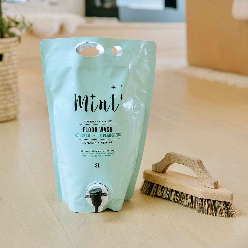 Mint Floor Wash eco-friendly refill pouch next to a wooden floor brush, set against a natural background. Ideal for environmentally conscious cleaning with a refreshing rosemary and mint scent.