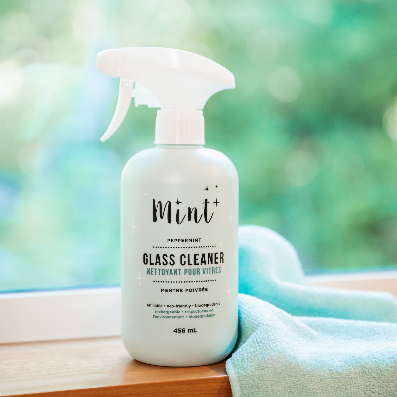 Mint Glass Cleaner bottle placed on a windowsill with a soft turquoise cloth beside it, evoking freshness and cleanliness.