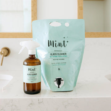 Mint Glass Cleaner in a 3L refill pouch with an easy-to-use pour spout, placed on a bathroom counter next to a 456ml glass spray bottle. The labels highlight its peppermint scent and natural ingredients.