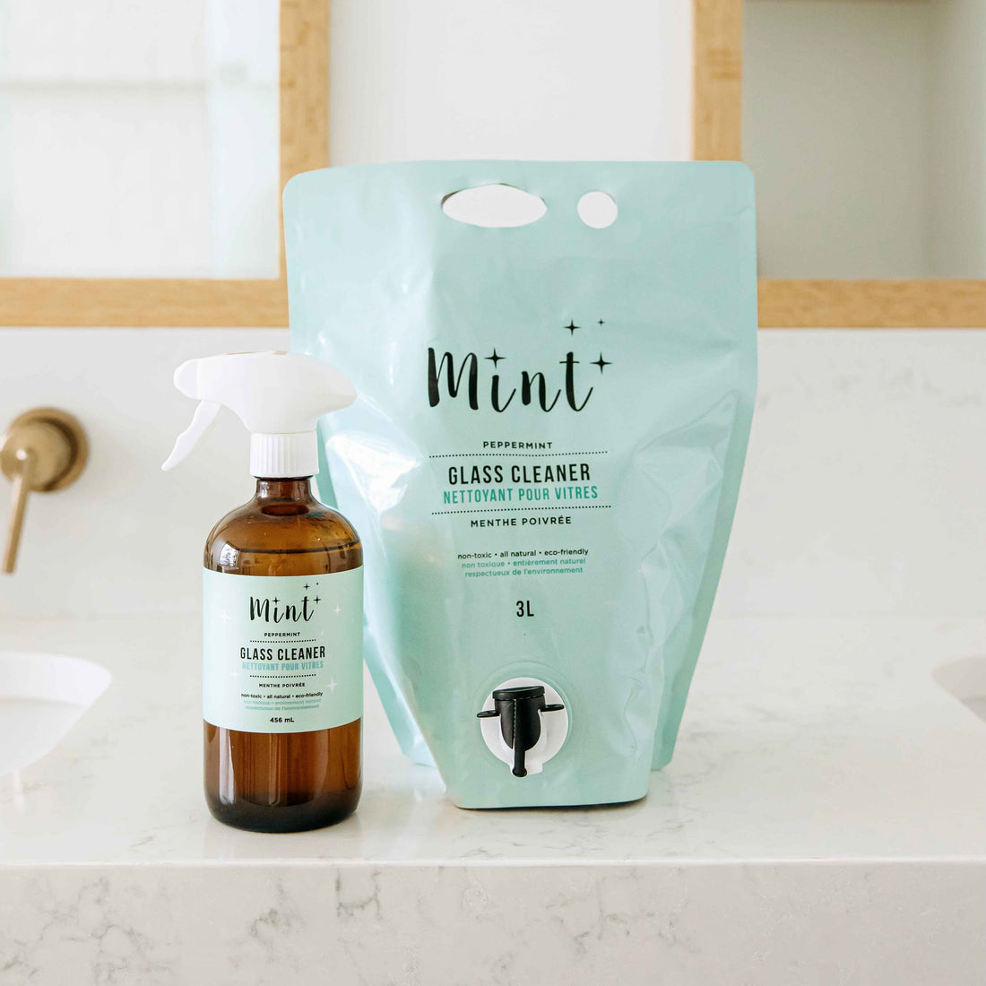 Mint Glass Cleaner in a refillable 3L eco-friendly pouch placed next to a glass spray bottle on a bathroom countertop with a marble surface.