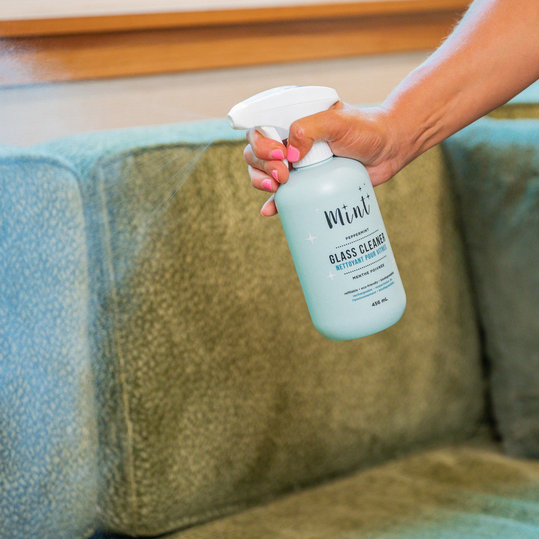 A person spraying Mint Glass Cleaner onto a green sofa to demonstrate its cleaning effectiveness, with a focus on the spray bottle in use.