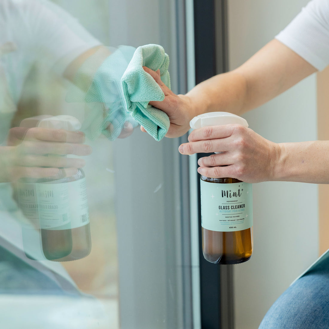 A person uses a Mint glass cleaner and a microfiber cloth to clean a window, demonstrating the product&