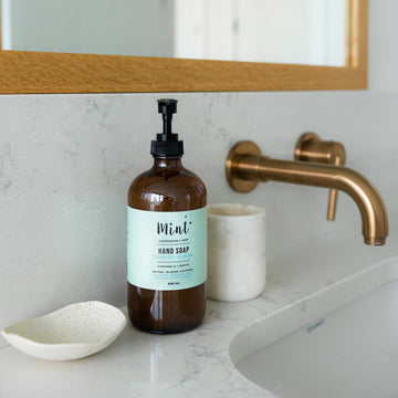 Mint Lemongrass & Mint Hand Soap in a 456ml eco-friendly glass bottle with a pump dispenser, placed on a bathroom counter next to a brass faucet, a ceramic soap dish, and a marble cup.