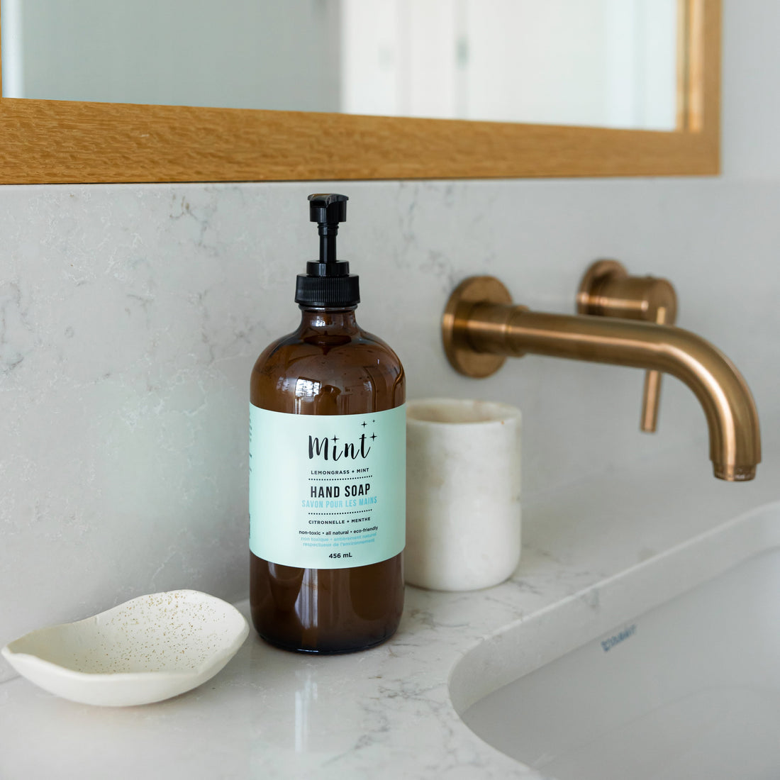 Mint Hand Soap in an amber glass bottle on a marble bathroom countertop beside a gold faucet, displaying its elegant and eco-conscious packaging.