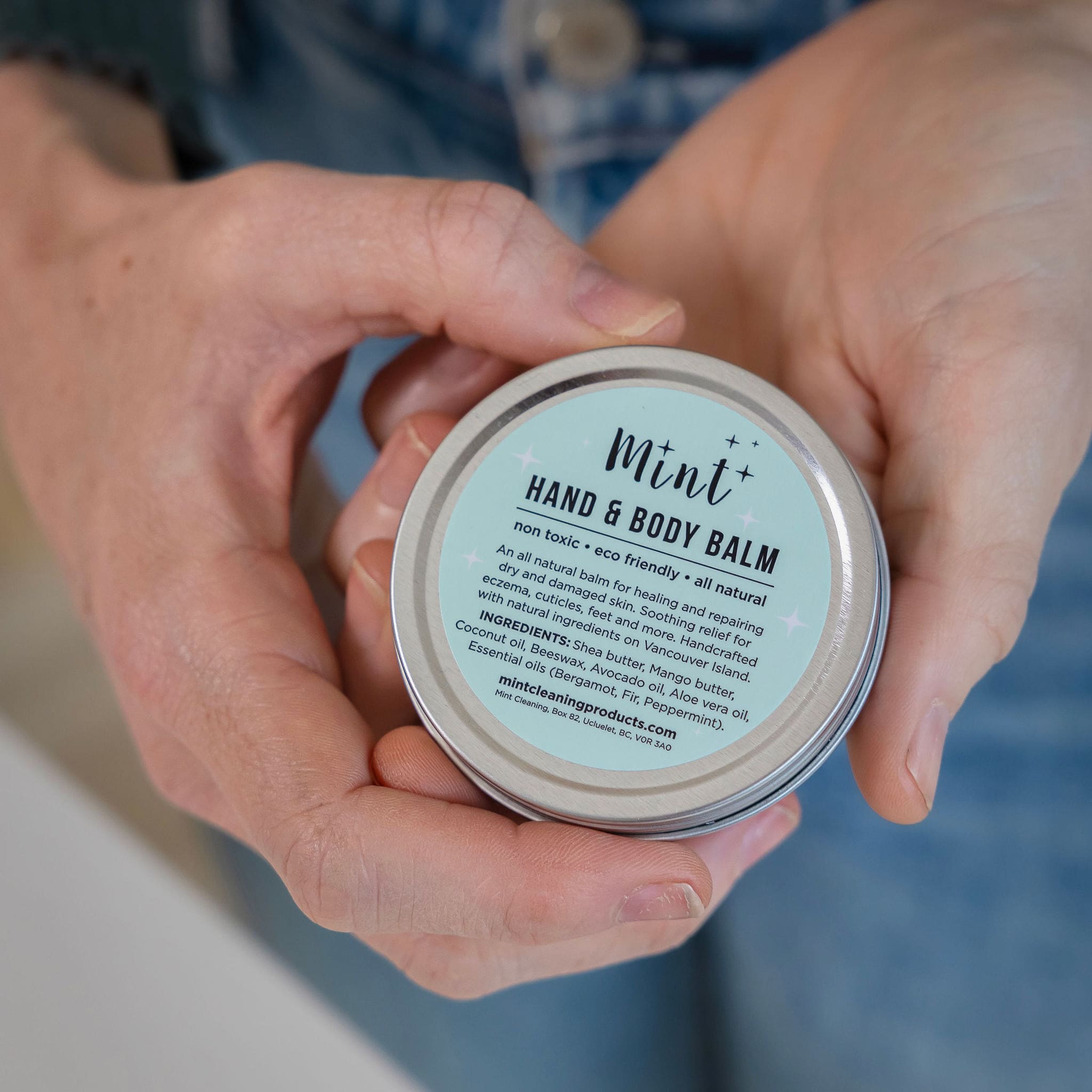 Person holding a tin of Mint Hand &amp; Body Balm, displaying the label with details about the product’s ingredients and benefits for dry, sensitive skin.