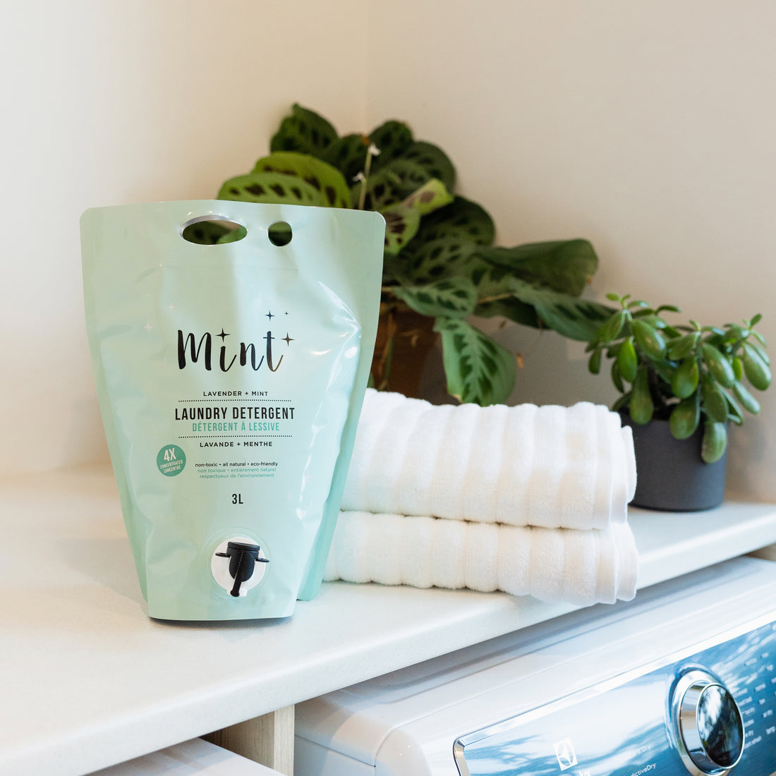 Mint Lavender & Mint Laundry Detergent in a 3L refill pouch with a spout, displayed on a laundry room counter next to folded white towels and a potted plant. The pouch highlights its natural, non-toxic, and eco-friendly ingredients.