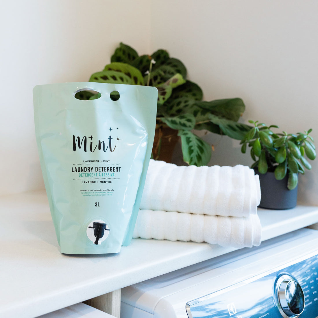 Eco-friendly Mint Laundry Detergent refill pouch in lavender and mint scent, displayed next to neatly folded towels and indoor plants in a laundry room setting.