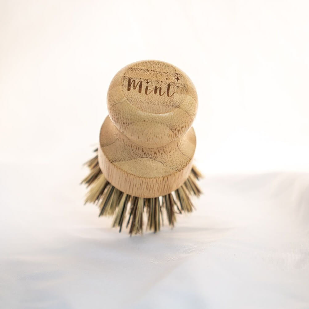 A close-up of a Mint bamboo dish brush with natural bristles, featuring the Mint logo engraved on the wooden handle, placed on a white surface.