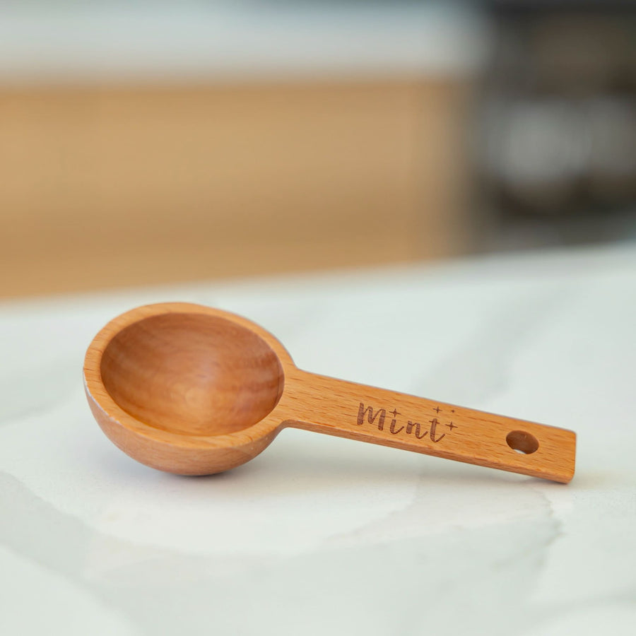 Close-up of Mint-branded wooden scoop with a smooth finish, resting on a white marble countertop.