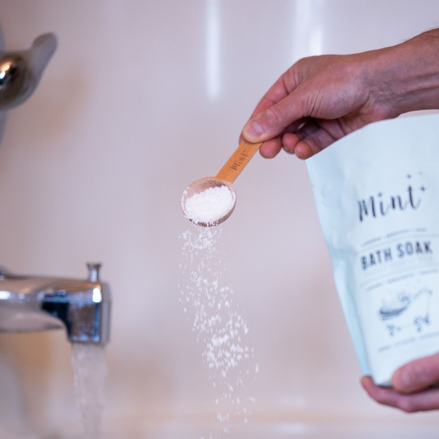 Hand holding a wooden scoop pouring Mint Bath Soak powder into a running bathtub for a relaxing soak experience.